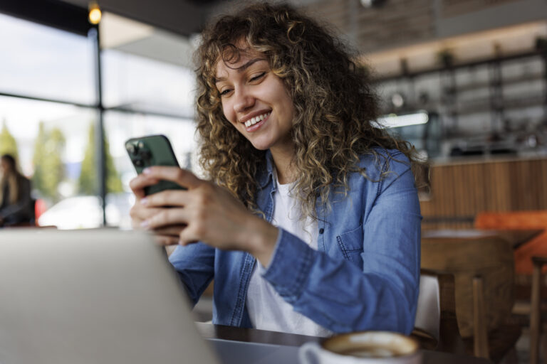 Young smiling woman using mobile phone while working on a laptop at a cafe