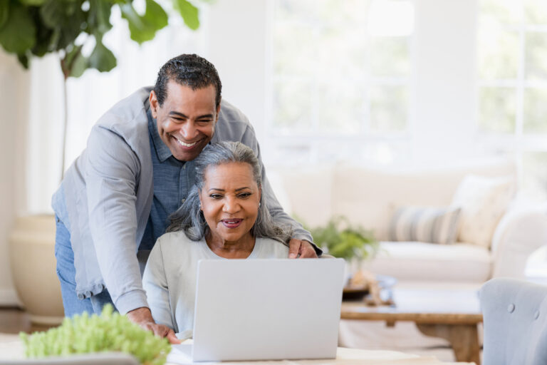 Senior husband looks over wife's shoulder at laptop screen