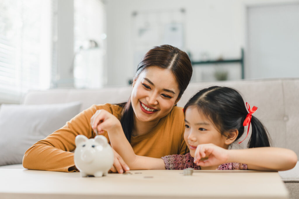 Cute little asian girl saving money by adding a coin in piggy bank with mother at home