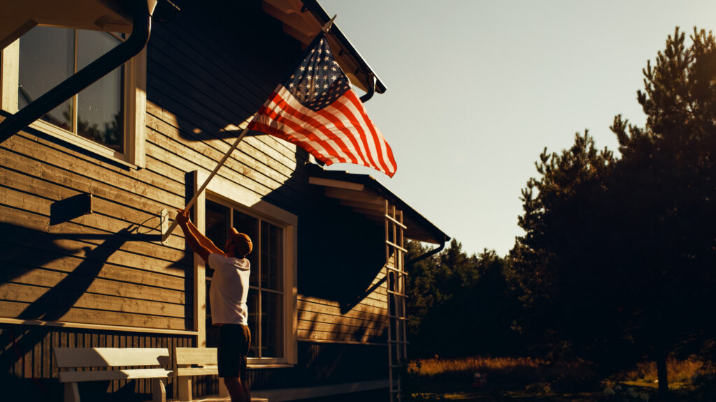 Proud American Homeowner Flying the United States of America National Flag on His Residential Area Home. Adult Man Fixing the Flag on His House Wall. Perfect Summer Day to Celebrate Patriotism.